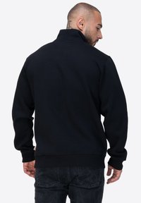 Felpa nera con colletto alto e polsini a coste. Realizzata in un tessuto morbido, presenta un'etichetta visibile sul lato inferiore ed è abbinata a jeans scuri.