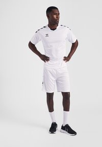 Weißes Sporttrikot und Shorts mit schwarzen Schulterakzenten. Das Modell trägt schwarze Sneakers und weiße Socken. Einfaches, leichtes Design.