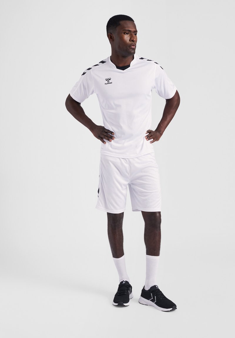 Hvid atletisk trøje og shorts med sorte skulderdetaljer. Modellen bærer sorte sneakers og hvide sokker. Enkel, letvægtsdesign.