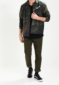 Giacca con motivo camouflage e bottoni a pressione, t-shirt nera, pantaloni verde oliva e sneakers nere con suole bianche e fibbie.