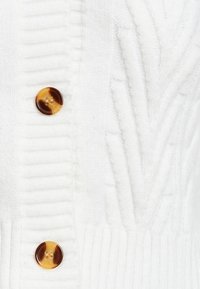 Cardigan en tricot blanc avec un design en rib et texturé, agrémenté de deux grands boutons ambre-brun avec un motif tourbillonnant.