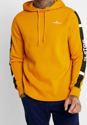 Homme portant un sweat à capuche G-Star Raw jaune moutarde avec des détails à rayures noires et blanches sur les manches et un pantalon bleu foncé, mains partiellement visibles.