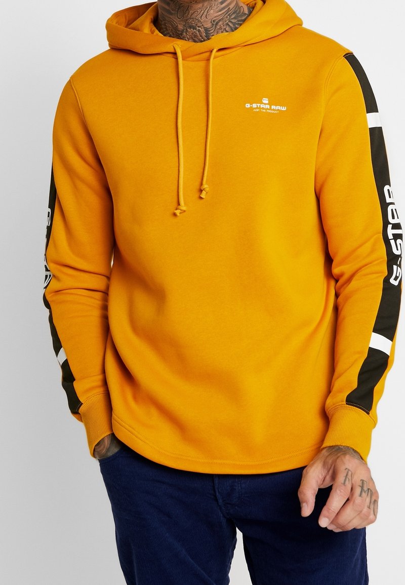 Homme portant un sweat à capuche G-Star Raw jaune moutarde avec des détails à rayures noires et blanches sur les manches et un pantalon bleu foncé, mains partiellement visibles.
