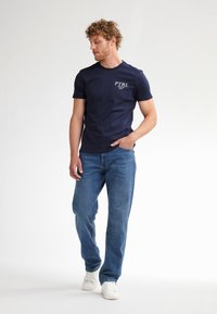 Marineblå bomuld t-shirt med korte ærmer og rund hals, med hvid "PTRL" logo på brystet. Kombineret med lyseblå jeans og hvide sneakers.