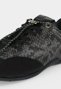 Gros plan d'une basket grise et noire avec une texture peau de serpent, lacets noirs, un verrou métallique pour lacets, et une semelle en caoutchouc arrondie.