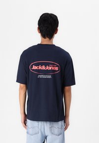 Tumši zils kokvilnas T-krekls ar īsām piedurknēm. Aizmugurē ir sarkans un balts teksts: "Jack & Jones" un "Designed in Denmark" apļa dizainā.