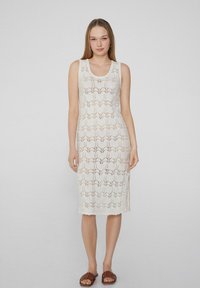 Weißes gestricktes Kleid mit floralem Spitzenmuster, ärmellosem Design, runder Halslinie und seitlichem Schlitz, kombiniert mit braunen Rutschsandalen.