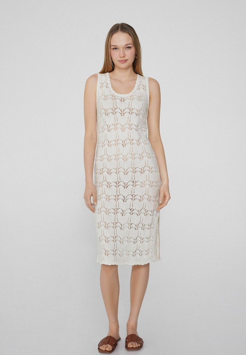 Weißes gestricktes Kleid mit floralem Spitzenmuster, ärmellosem Design, runder Halslinie und seitlichem Schlitz, kombiniert mit braunen Rutschsandalen.