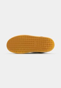 Semelle d'une sneaker noire avec un fond en caoutchouc jaune texturé, présentant un motif en grille et un logo subtil au centre.