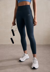Magas derekú, sötétkék leggings, texturált mintával, fehér sportcipővel párosítva. Az egyik kezében ugrókötél van.