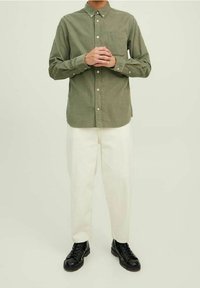 Camicia verde oliva con bottoni e taschino sul petto, abbinata a pantaloni bianchi e scarpe nere, con una texture semplice e vestibilità standard.