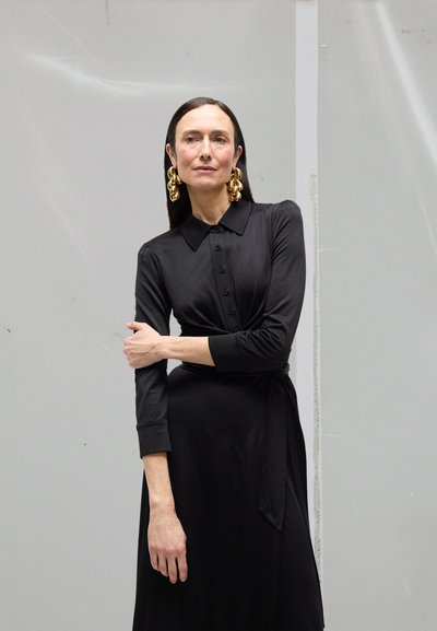 Vestido cruzado negro con cuello, mangas largas y detalles de botones. Pendientes dorados destacados y textura suave. Diseño simple y elegante.