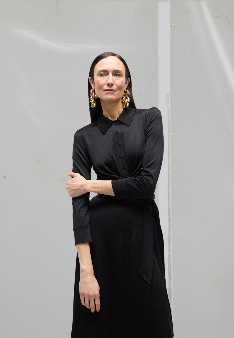 Rochie petrecută neagră cu guler, mâneci lungi și detalii cu nasturi. Cercei aurii remarcabili și textură netedă. Design simplu și elegant.