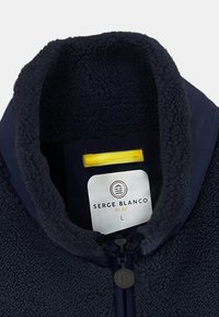 Veste en polaire navy avec une texture douce, un col haut et une fermeture éclair. L'étiquette indique "SERGE BLANCO" avec un accent jaune au-dessus. Taille L.