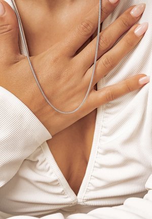 Eine silberne Kettenhalskette liegt über einer Hand und zeigt ein glattes, elegantes Design. Der strukturierte weiße Stoff der Kleidung steht im Kontrast zur Haut.