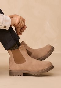 Beige ruskind ankelstøvler med elastiske sider og en robust gummisål. Designet har en syningsdetalje og en lav hæl.