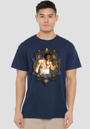 Young man wearing navy t-shirt with graphic of Elvis Presley singing and text "Aloha from Hawaii" standing against plain background.