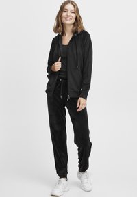Survêtement en velours noir comprenant un hoodie zippé avec une poche avant et un pantalon assorti à taille élastique. Des baskets blanches viennent parfaire le look.