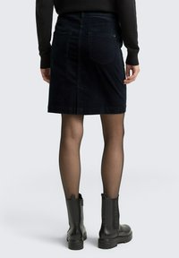 Jupe en denim foncé avec une silhouette ajustée, deux poches arrière et une fente à l'arrière. Portée avec des bottines noires et des collants noirs transparents.