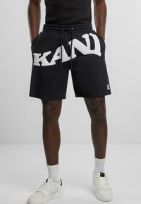 Black shorts made of cotton, featuring a white fluffy "KAND" logo on the front, with an elastic waistband and side pockets.
