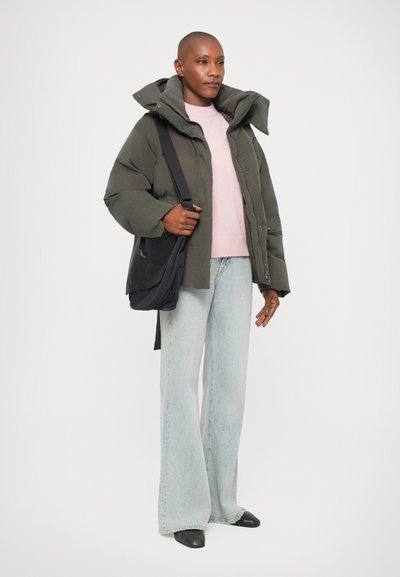 Persoon in een groene pufferjack over een roze trui, lichtblauwe wijde jeans, zwarte schoenen en met een zwarte schoudertas.
