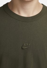 Oliwkowy bawełniany T-shirt z okrągłym dekoltem. Z przodu po lewej stronie piersi znajduje się haftowane logo Nike w tym samym kolorze.