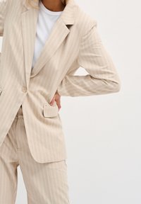 Béžový károvaný blazer s jedním knoflíkem a předními kapsami, v kombinaci s odpovídajícími kalhotami, vyrobený z lehké tkaniny.