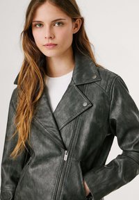 Tume roheline kunstnahast mootorrattajope, millel on krae, diagonaalne tõmblukk, tikitud õlavarre aktsendid ja küljetaskud.