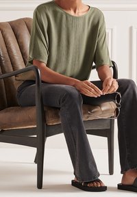 Top corto a maniche corte verde oliva con una vestibilità ampia, abbinato a jeans svasati scuri e sandali neri flat. Seduto su una sedia marrone con una texture.