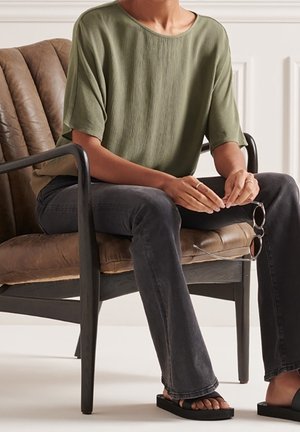 Top corto a maniche corte verde oliva con una vestibilità ampia, abbinato a jeans svasati scuri e sandali neri flat. Seduto su una sedia marrone con una texture.