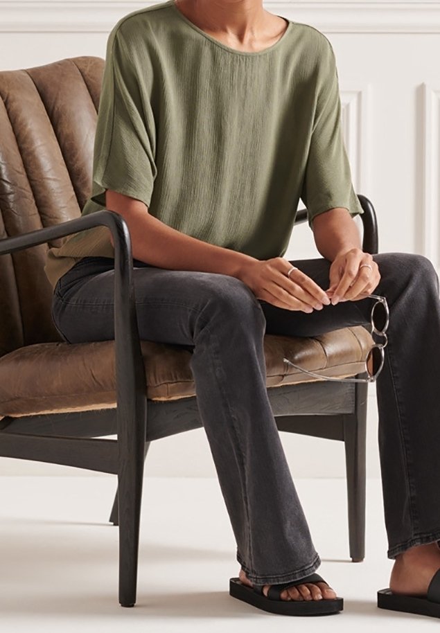 Top corto a maniche corte verde oliva con una vestibilità ampia, abbinato a jeans svasati scuri e sandali neri flat. Seduto su una sedia marrone con una texture.