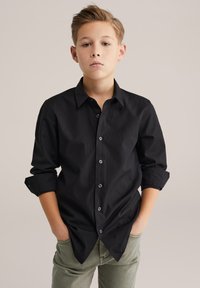 Melna kokvilnas pogājama krekls ar klasisku apkakli, garām piedurknēm, kas uzlocītas manšetēs, un priekšējām kabatām. Valkāts ar olīvu zaļām biksēm.