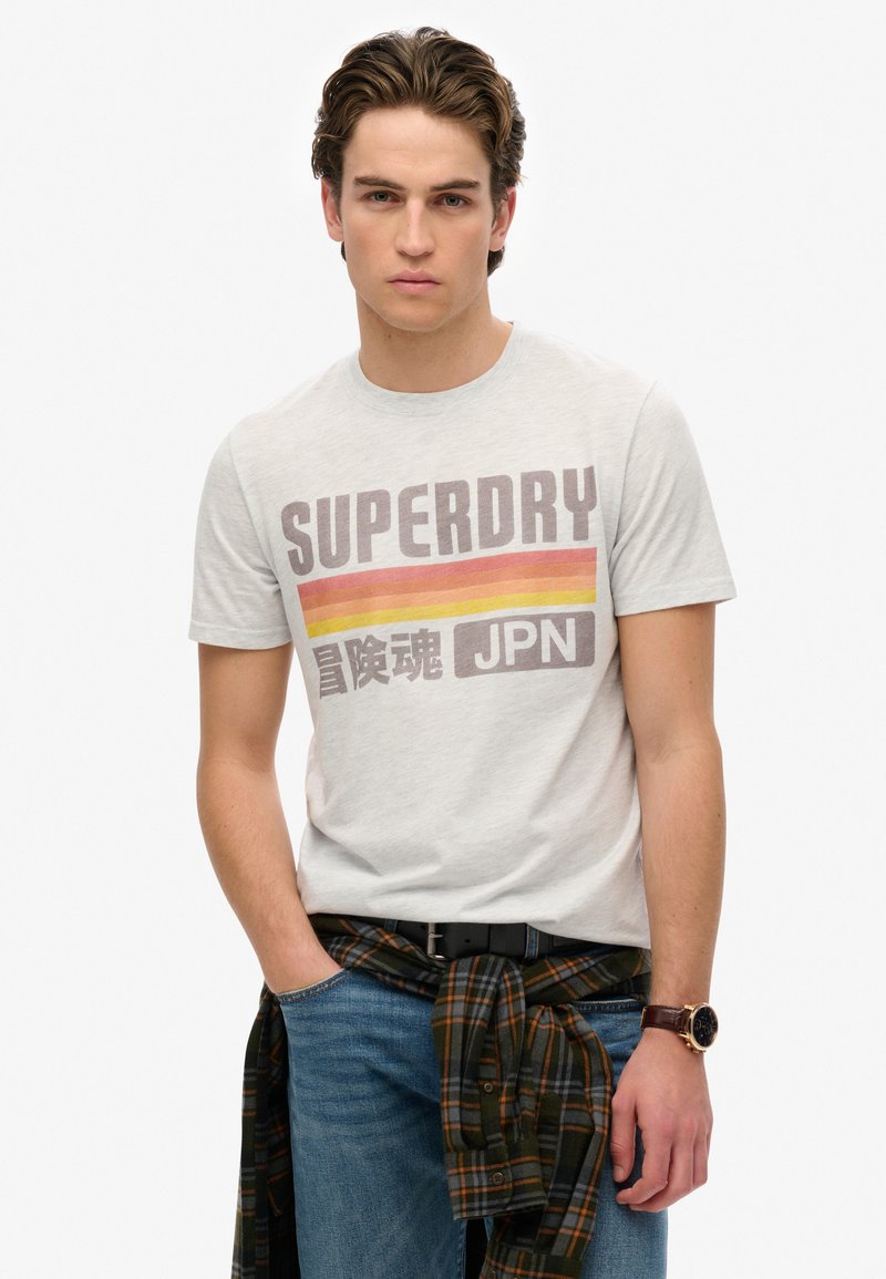 Camiseta de algodón gris claro con el texto "SUPERDRY" en rayas marrones y naranjas. Llevada con jeans azules y una camisa de cuadros verde atada a la cintura.