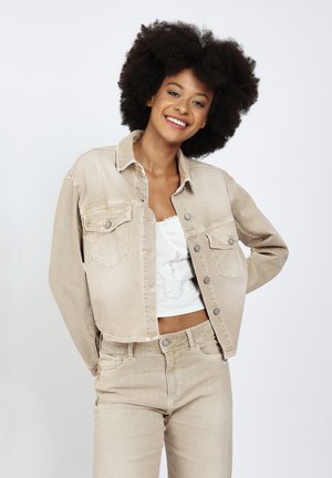 Junge Frau mit natürlichem Afro-Haar, die lächelt, trägt eine beige Jeansjacke und Jeans mit weißem Spitzentop, steht vor einem schlichten Hintergrund.