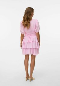 Rosa, lager-på-lager klänning med korta puffärmar, smockad midja och textil med struktur; bärs med beiga skor, sedd från baksidan.