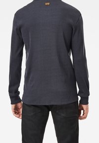 Marinblå långärmad T-shirt, ribbad textur, rund halsringning och G-Star-märke i nacken. Matcher med mörka byxor, händer synliga.