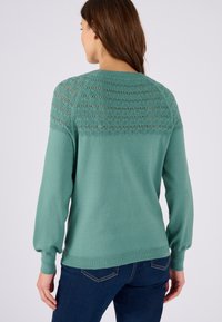 Pull vert avec un tricot texturé en maille ouverte au niveau des épaules, manches longues bouffantes et poignets côtelés. Associé à un jean bleu foncé.