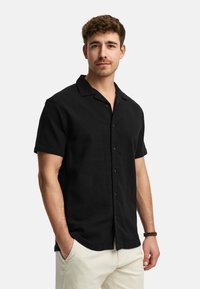 Black short-sleeve button-up shirt made of textured fabric, featuring a camp collar and visible buttons, paired with light-colored pants.