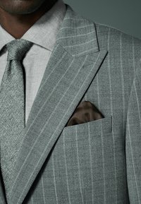 Traje gris a rayas con un acabado texturizado, combinado con una camisa gris clara y una corbata a juego. Pañuelo de bolsillo marrón visible en el bolsillo de la chaqueta.