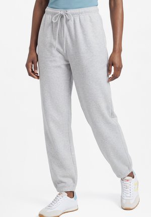 Persoon draagt lichtgrijze joggingbroek met elastische boorden en witte sneakers met blauwe en gele accenten, staand tegen een effen achtergrond.