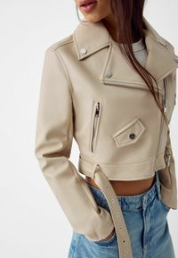 Giacca motociclista corta in ecopelle beige con colletto a revers, tasca con zip, dettagli con bottoni a pressione e vita con cintura.