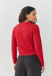 Red, textured sweater with long sleeves and a round neckline. Cropped design; worn with dark, tailored pants. Simple, clean lines.