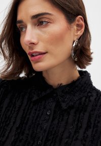 Black textured shirt with vertical fringing, featuring a collar and buttons. Silver statement earrings add a reflective detail.
