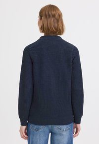 Pull en maille côtelée bleu marine avec manches longues, col et un dos légèrement plus long. Tissu léger avec texture.