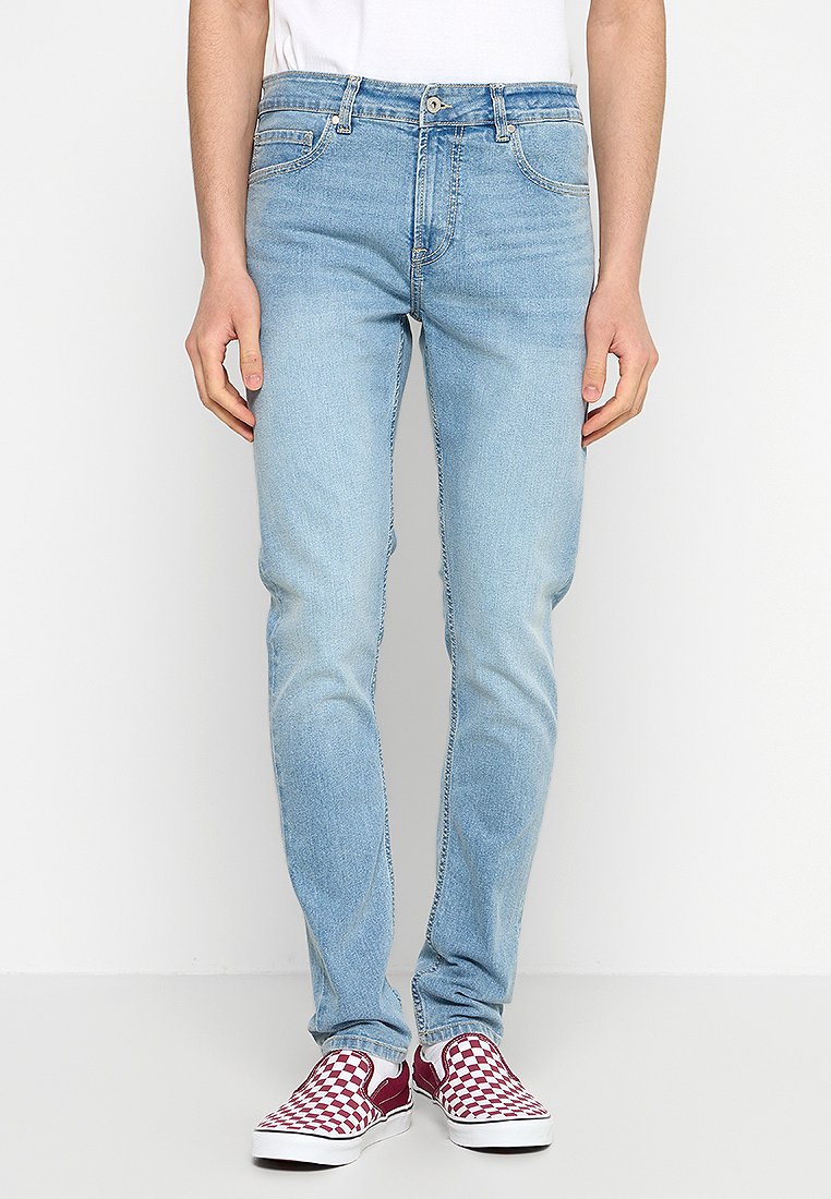 Vaqueros de denim azul claro con un corte ajustado, que presentan un diseño clásico de cinco bolsillos y un ligero desgastado. Combinados con zapatos tipo slip-on de estampado a cuadros burdeos.