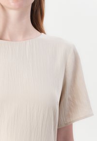 Beige kortermet topp med rynkete tekstur, rund hals og avslappet passform. Laget av lett stoff med minimale sømmer.