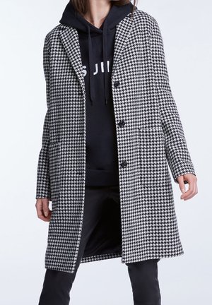 Femme portant un manteau à motif pied-de-poule noir et blanc par-dessus un sweat à capuche noir et un pantalon noir, debout devant un fond uni.