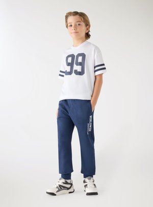 Ragazzo in piedi con le mani in tasca, indossa una maglietta bianca con il numero 99 e pantaloni della tuta blu con la scritta "Worldwide" e scarpe da ginnastica bianche.