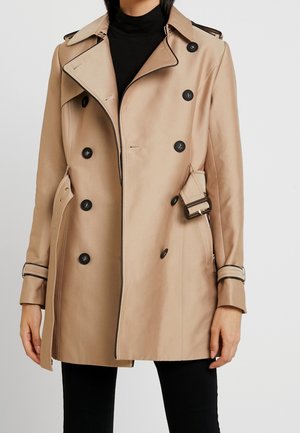 Person trägt einen beigen, zweireihigen Trenchcoat mit schwarzen Knöpfen und Gürtel über einem schwarzen Rollkragenpullover und Hose.