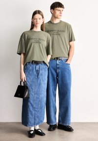 Tricouri de bumbac verde măsliniu cu imprimeu "TOMMY JEANS", asortate cu denim albastru: femeie în fustă lungă, bărbat în blugi largi, amândoi cu curele.